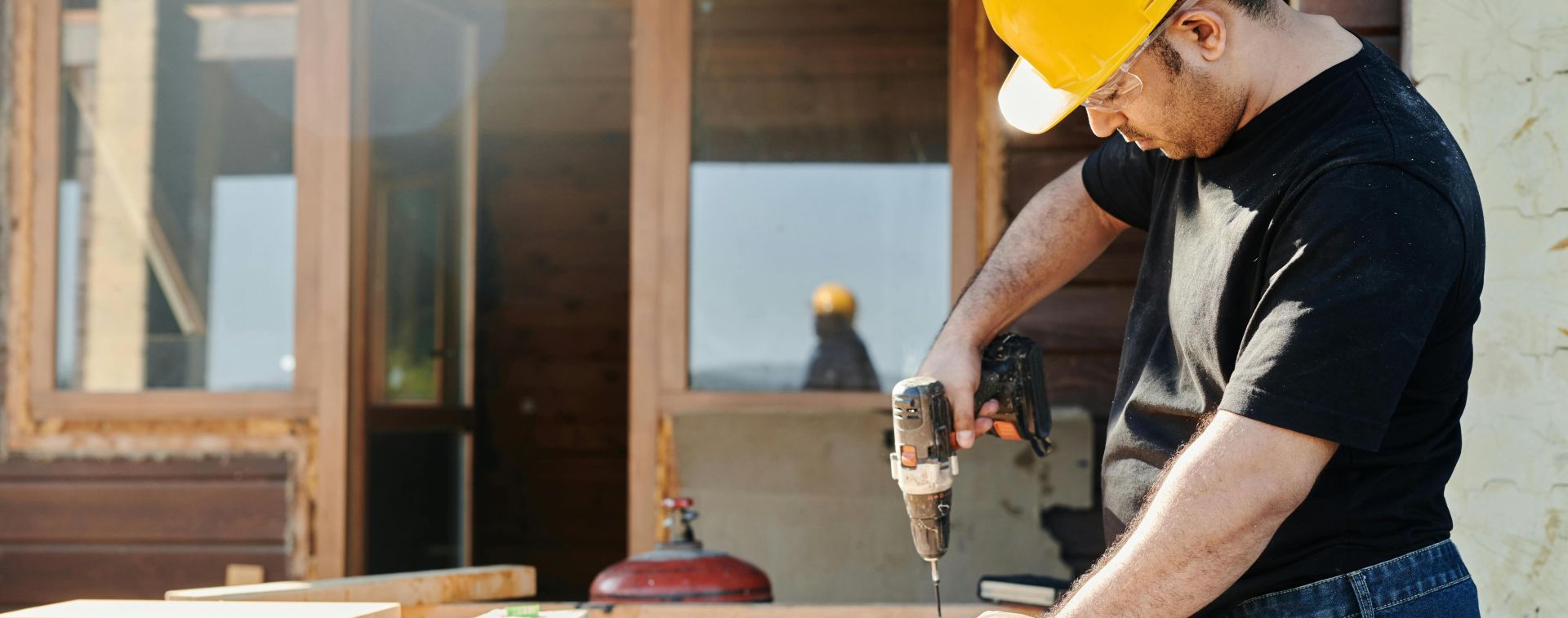 A construction worker with a hard hat uses a power drill on a wooden plank at a renovation site.
