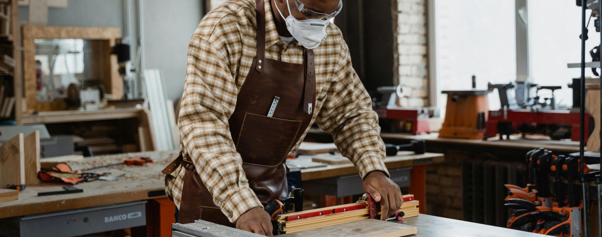 Artisan wearing an apron and mask, precisely measuring and cutting wood in a workshop setting.