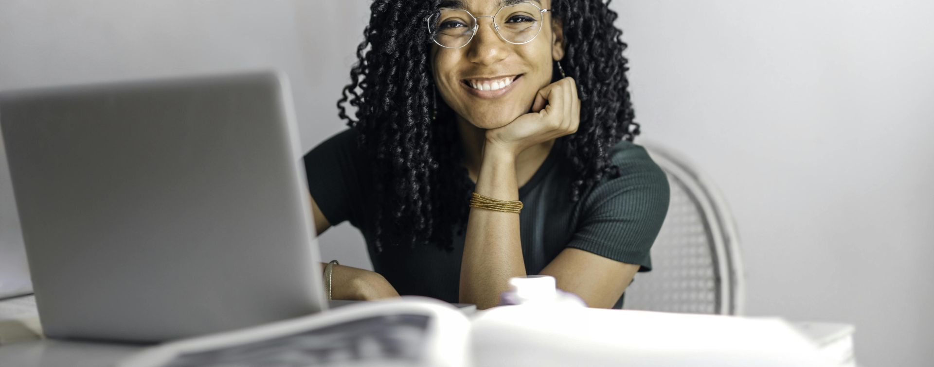 Joyful businesswoman with curly hair smiling at camera while using laptop indoors.